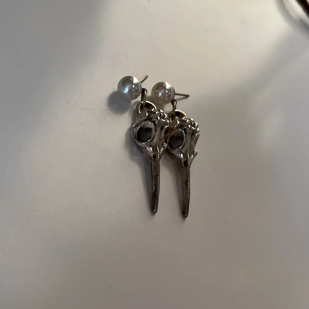 Unika örhängen i silverfärg med en pärldetalj vid fästet. Designen har en intressant form som påminner om en fågelnäbb, vilket ger en edgy och stilren look. Perfekta för att ge en extra touch till din outfit.. Asusteet.