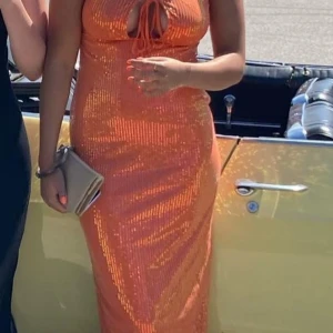 Orange glittrig klänning - Säljer en fantastisk orange långklänning med glittriga paljetter. Klänningen har en v-ringad design med knytning framtill och är ärmlös. Hade på mig den på min bal fick många komplimanger!