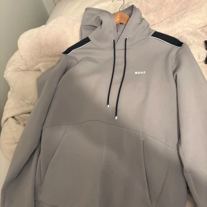 Grå hoodie från Boss - Snygg grå hoodie från Boss med svart detalj på axlarna och justerbar huva. Perfekt för en avslappnad stil. Den har en klassisk känguruficka framtill och diskret logga på bröstet.