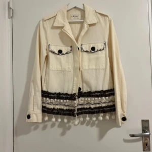 Beige jacka med dekorativa detaljer från Robin - Snygg beige jacka från Robin med unika dekorativa detaljer. Jackan har långa ärmar och knappar framtill. Två bröstfickor med svarta knappar och broderade kanter. Nedre delen pryds av svarta och vita mönster samt hängande tofsar.