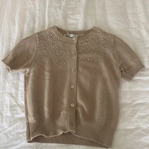 Beige kofta från Zara med pärlor - Säljer en söt beige kofta från Zara med korta ärmar och fina pärldetaljer på framsidan. Koftan har knappar framtill och ribbade kanter vid ärmslut och nederkant. Perfekt för en stilren look!