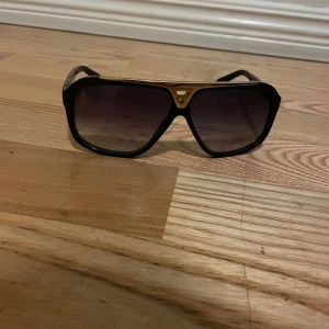 Lv  solglasögon med gulddetaljer - Lv  solglasögon med breda bågar och gulddetaljer på ovansidan. De har en klassisk design med mörka glas som ger en cool look. Perfekta för soliga dagar och att ge en touch av stil till din outfit.