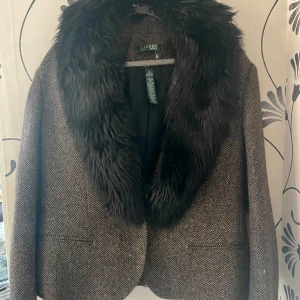 Ralph Lauren Brun kavaj med päls - Snygg brun kavaj med en lyxig fuskpälsdetalj runt kragen från Ralph Lauren. Perfekt för att ge en elegant touch till din outfit. Kavajen har en klassisk passform och är tillverkad i ett stilrent mönster.