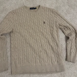 Beige stickad tröja från Ralph Lauren - Säljer en stilren beige stickad tröja från Ralph Lauren med kabelstickat mönster och broderad logotyp på bröstet. Tröjan har långa ärmar och rund halsringning. Den är varm och ger dig en old money look samt Perfekt för kyligare dagar! Inga defekter. Storlek S men passar både s-m, riktigt stretchigt material som gör att den passar på nästan alla 
