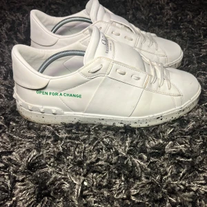 Vita sneakers från Valentino - Snygga vita sneakers från Valentino med texten 'OPEN FOR A CHANGE' i grönt på sidan. Skorna har en stilren design med snörning och en mönstrad sula. Perfekta för en trendig look. Box ingår.