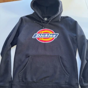 Svart hoodie från Dickies - Säljer en svart hoodie från Dickies med deras ikoniska logga i rött och gult på bröstet. Tröjan har en klassisk känguruficka och justerbar huva med dragsko. Perfekt för en avslappnad stil.