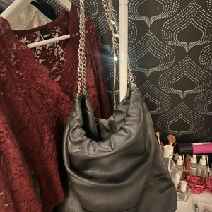 Svart axelväska med kedjedetaljer - Snygg svart axelväska med kedjedetaljer som ger en cool touch. Perfekt för att bära dina dagliga nödvändigheter med stil. Väskan har en mjuk och slouchy design som gör den både trendig och praktisk. Nypris 400-500 kr💗