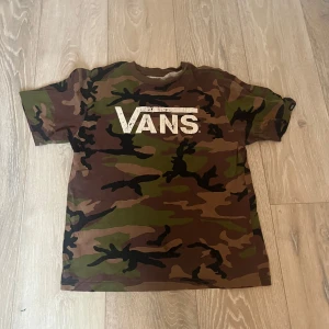 Camouflage t-shirt från Vans - Säljer en cool camouflage t-shirt från Vans i storlek L. T-shirten har ett stort Vans-tryck på framsidan och är tillverkad i 100% bomull. Perfekt för en avslappnad stil.