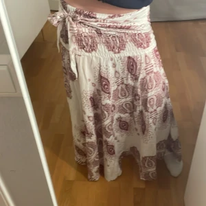 Vit och vinröd långkjol med mönster - Säljer en vacker långkjol i vitt och vinrött med ett detaljerat mönster. Kjolen har en knytning i midjan som ger en fin passform. 