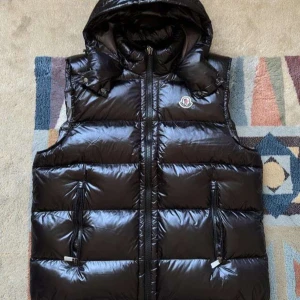 Svart väst från Moncler - Snygg svart väst från Moncler med glansig yta och huva. Västen har dragkedja och två fickor framtill. Perfekt för kyligare dagar.