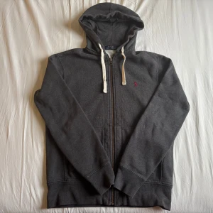 Grå hoodie från Polo Ralph Lauren - Säljer en stilren mörkgrå zip hoodie från Ralph lauren. Skicket är så gott som ny, inget täcken på användning. Storlek S men passar även medium. Skriv vid funderingar!