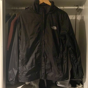 Svart vindjacka från The North Face - Säljer en svart vindjacka från The North Face i bra skick. Jackan är perfekt för regniga dagar med sitt vattentäta material och andningsfunktion. Den har en dragkedja framtill och en justerbar huva. Passar perfekt för höst- och vårsäsongen.