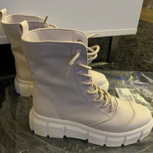 Beige boots med snörning - Säljer ett par snygga beige boots i syntetmaterial med rejäl snörning och en chunky sula. Perfekta för höst och vinter med sin robusta design. De är i nytt skick och har en modern look som passar till många outfits. Storleken är dam 38. 