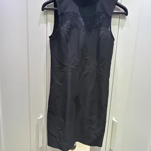 Svart ärmlös klänning med spetsdetalj - Snygg svart ärmlös klänning i mycket bra skick. Den har en elegant spetsdetalj över bröstet som ger en fin touch. Perfekt för festliga tillfällen eller en utekväll. Klänningen är knälång och har en figurnära passform. Använd bara en gång!