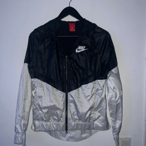 Svart och silver vindjacka från Nike - Säljer en snygg svart och silverfärgad vindjacka från Nike i storlek S. Jackan har en dragkedja framtill och en huva, perfekt för blåsiga dagar. Den är i bra skick och passar både till träning och vardagsbruk. Perfekt för vår och höst!
