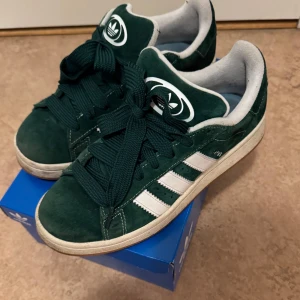 Gröna Adidas Campus sneakers - Säljer ett par snygga gröna Adidas Campus sneakers i mocka. De har de klassiska vita ränderna på sidan och en bekväm passform. Perfekta för vardagsbruk och ger en stilren look. Skorna är i bra skick med lite slitage på sulan. Kommer i originalkartong.
