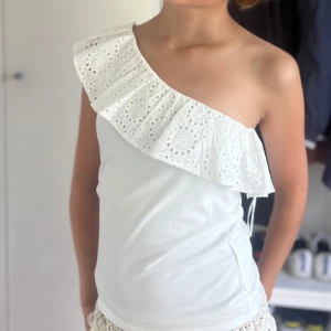 Vit oneshoulder topp med volang - Säljer en superfin vit oneshoulder topp med volangdetalj. Perfekt för sommardagar eller festliga tillfällen. Toppen är i ett mjukt och bekvämt material som sitter skönt mot huden. Passar perfekt till både jeans och kjol! Köpt på Lindex! Ny skick strl S endast använd 1 gg