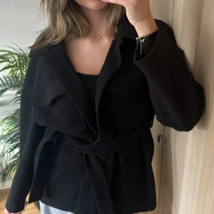 Svart kappa med bälte - Säljer en stilren svart kappa med knappar och bälte i midjan. Perfekt för höst och vår, med en normal passform och långa ärmar. Kappan är i mycket bra skick och passar både till vardags och fest. Från gina