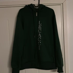 Mörkgrön hoodie med strassdetaljer - Säljer en snygg mörkgrön hoodie med dragkedja från Lecacci. Den har coola strassdetaljer med text på framsidan och en fjäril på baksidan. Perfekt för höst och vinter när du vill vara både varm och stilren. Passar perfekt till vardags eller en chill kväll med vänner. Tror att nypris ligger runt 900kr. Pris kan diskuteras!😊