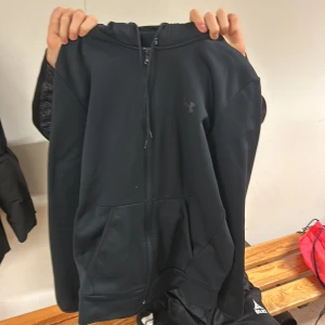 Svart hoodie från Under Armour - Säljer en svart hoodie från Under Armour i storlek S. Den är i bra skick och har en dragkedja framtill. Perfekt för träning eller vardagsbruk med en bekväm och lös passform. Den har en klassisk design med fickor framtill och en mjuk insida för extra komfort.