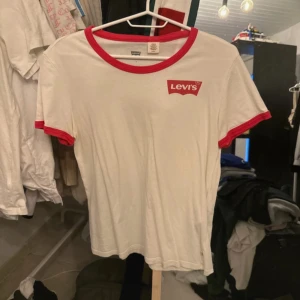 Vit och röd t-shirt från Levi's - Säljer en snygg vit t-shirt från Levi's med röda detaljer. Den har en röd krage och röda kanter på ärmarna. Perfekt för en casual look eller en dag ute med vänner. T-shirten är i bomull och har en normal passform.