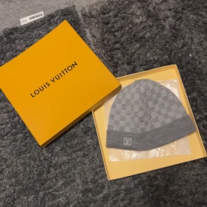 Grå rutig mössa från Louis Vuitton - Säljer en snygg grå mössa från Louis Vuitton med ett klassiskt rutigt mönster. Mössan är i mycket bra skick och har en broderad LV-logga framtill. Perfekt för att hålla sig varm med stil under höst och vinter. Kommer i originalförpackning.