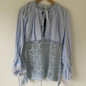 Ljusblå blus från Hunkydory - Säljer en superfin ljusblå blus från Hunkydory. Den har långa ärmar med knytband och vackra spetsdetaljer. Blusen är i ett luftigt material med en unik design där nederdelen är i spets. Perfekt för vår och sommar!