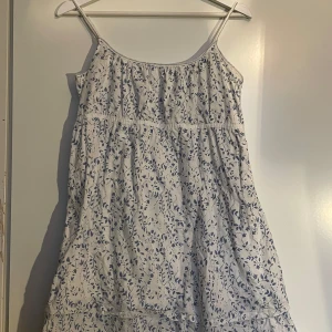 Vit blommig sommarklänning - Söt vit klänning med blått blommönster, perfekt för sommardagar. Den är ärmlös med smala axelband och har en luftig passform. Klänningen är i mycket bra skick och en kortare passform!