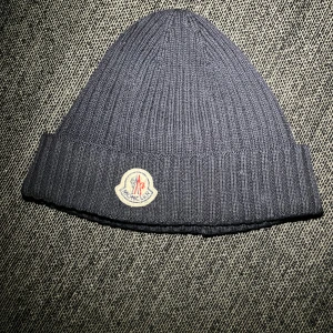 Moncler mössa - Säljer en snygg mörkblå ribbad mössa från Moncler. Den är perfekt för kalla dagar och har en broderad Moncler-logga framtill. Mössan är i mycket bra skick och passar perfekt till både vardag och vinteraktiviteter. Håll dig varm och stilren! 🧢❄️priset kan sänkas vid snabb affär