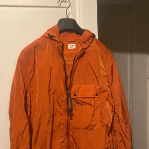 Orange jacka från C.P. Company - Säljer en cool orange jacka från C.P. Company. Den har en unik design med inbyggda glasögon i huvan och en praktisk dragkedja framtill. Perfekt för höst och vår, och den är i jätte bra skick . Jackan är gjord av syntetmaterial och har en normal passform.