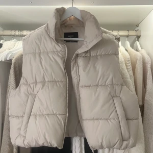 Beige dunväst från Bik Bok - Säljer en snygg beige dunväst från Bik Bok i storlek M. Västen har en dragkedja framtill och är perfekt för höst och vår. Den är i mycket bra skick och gjord av återvunnet material. Perfekt för att hålla sig varm och stilren!