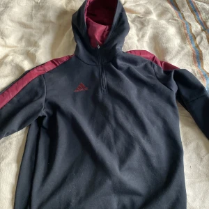 Svart och vinröd hoodie från Adidas - Säljer en snygg svart hoodie från Adidas med vinröda detaljer. Den har en halv dragkedja framtill och klassiska tre ränder längs ärmarna. Perfekt för höst och vinter, och den är i bra skick. Passar både för träning och vardagsbruk.