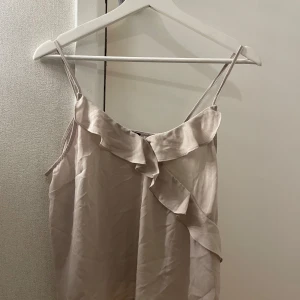 Beige linne med volangdetaljer - Säljer ett stilrent beige linne med smala axelband och fina volangdetaljer framtill. Perfekt för sommardagar eller en avslappnad kvällslook. Materialet är lätt och luftigt, vilket gör det bekvämt att bära.