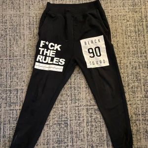 Svarta mjukisbyxor med tryck - Säljer ett par svarta mjukisbyxor från FSBN i storlek S. Byxorna har coola vita tryck med texten 'F*CK THE RULES' och 'BLACK 90 SQUAD'. Perfekta för en avslappnad stil eller chill hemma. De är i bra skick och gjorda av bomull för extra komfort.