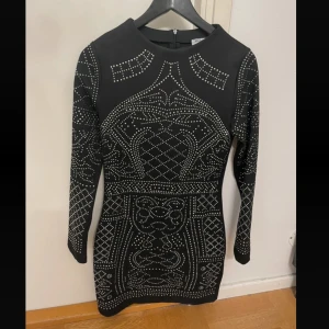 Svart klänning med paljetter - Snygg svart klänning från Boohoo i storlek S. Den har ett coolt mönster med glittriga detaljer och långa ärmar. Perfekt för en festkväll! Klänningen är tight och har en dragkedja i ryggen för enkel påklädning.