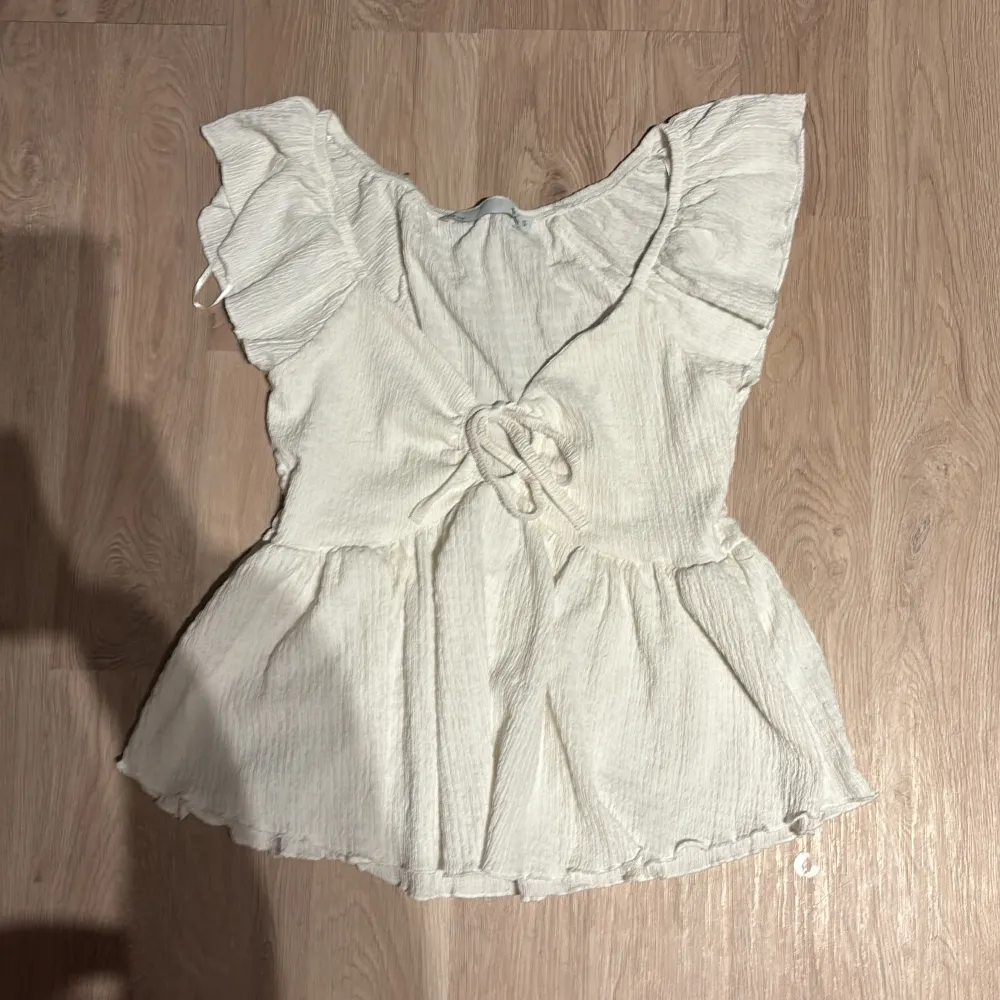 Söt vit blus med volangdetaljer och knytning framtill. Perfekt för sommardagar eller en avslappnad vårlook. Den har en luftig passform och är tillverkad i ett lätt material som ger en romantisk känsla.. Yläosat.