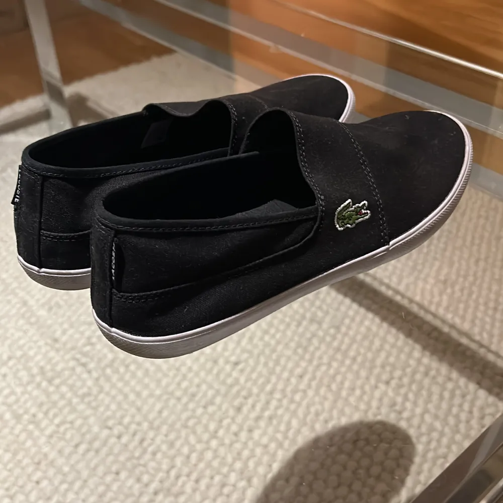 Säljer ett par stilrena svarta slip-on sneakers från Lacoste. De har en enkel design med den klassiska krokodilloggan på sidan. Perfekta för en avslappnad stil och lätta att ta på. Sulan är vit och ger en snygg kontrast till det svarta tyget.. Skor.