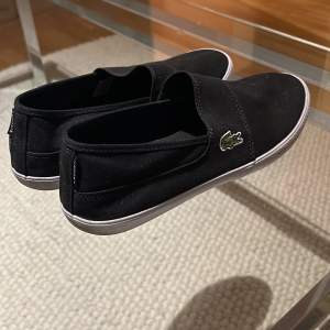 Säljer ett par stilrena svarta slip-on sneakers från Lacoste. De har en enkel design med den klassiska krokodilloggan på sidan. Perfekta för en avslappnad stil och lätta att ta på. Sulan är vit och ger en snygg kontrast till det svarta tyget.