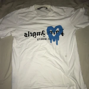 Vit t-shirt med blått hjärta - Säljer en vit t-shirt från Palm Angels med ett coolt blått hjärta och text på framsidan. Tyvärr finns det några små hål i tyget, men den är fortfarande snygg och perfekt för en avslappnad stil. Passar bra till jeans eller shorts!