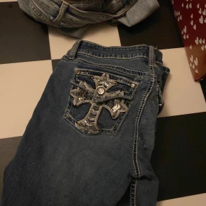 Blå jeans från Antique Rivet - Säljer ett par snygga blå jeans från Antique Rivet. De har en cool broderad detalj på bakfickan med en korsdesign och glittriga stenar. Jeansen är lagade en gång vid fickan där bak❤️