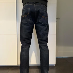 Mörkblå jeans med broderad detalj - Säljer ett par mörkblå jeans i mycket bra skick. De har en snygg broderad detalj på bakfickorna och en klassisk femficksdesign. Jeansen är av normal passform och har en midwaist. Perfekta för både vardag och fest!