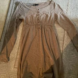 Brun  långärmad klänning från Cubus - Mysig beige klänning från Cubus i mjukt material. Den har långa ärmar och knappar framtill som ger en söt detalj. Perfekt för höst och vår, och funkar både till vardags och lite finare tillfällen. Passar bra med ett par leggings eller tights.