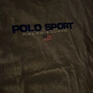 Grå t-shirt från Polo Sport - Säljer en grå t-shirt från Polo Sport av Ralph Lauren. Den har en klassisk design med korta ärmar och ett tryck med texten 'POLO SPORT' och en amerikansk flagga på framsidan. Perfekt för en avslappnad stil!