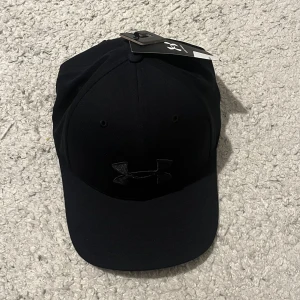 Under Armour keps - Säljer en snygg svart keps från Under Armour i nytt skick. Kepsen har en justerbar rem baktill för perfekt passform och är tillverkad i bomull. Perfekt för både vardag och träning!