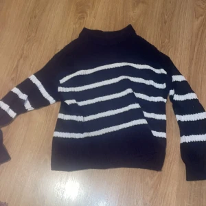 Marinblå och vit randig stickad tröja från 157 - Säljer en blå stickad tröja med vita ränder från 157. Den har en mysig och varm känsla, perfekt för kyliga dagar. Tröjan har långa ärmar och en klassisk passform. Perfekt för både vardag och avslappnade tillfällen, inte alls använd många gånger.  
