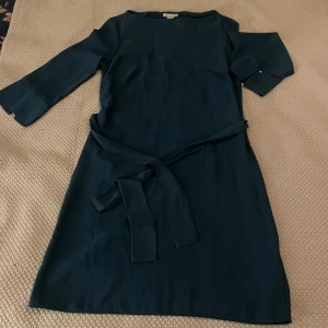 Mörkgrön klänning från H&M - Snygg mörkgrön klänning från H&M i storlek M. Den har trekvartsärmar och ett knytband i midjan för en fin passform. Perfekt för både vardag och festliga tillfällen. Materialet känns mjukt och bekvämt mot huden.
