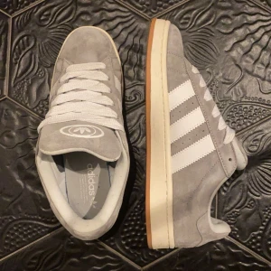 Grå Adidas Campus - Säljer ett par snygga grå Adidas Campus i mocka med vita detaljer och klassiska tre ränder. Skorna har en bekväm passform och en gummisula i ljusbrun färg. Perfekta för vardagsbruk och ger en stilren look. De är i mycket bra skick 10/10 som syns på bilderna. Tyvär ingen box då jag slängde den, men endast testade.