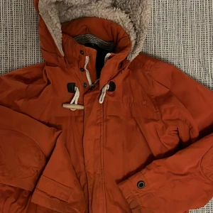Orange vinterjacka med foder strlk 164 - Säljer en varm och gosig orange vinterjacka med mjukt foder i luvan. Jackan har dragkedja och knappar framtill för extra skydd mot kylan. Perfekt för kalla vinterdagar! 🧡Strlk 164 