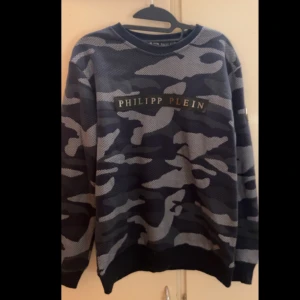 Camouflage tröja från Philipp Plein - Säljer en oanvänd tröja från Philipp Plein. Den har ett grått och svart mönster med märkets logga på bröstet. Tröjan är långärmad och perfekt för en avslappnad stil. Passar bra till både vardag och fest!
