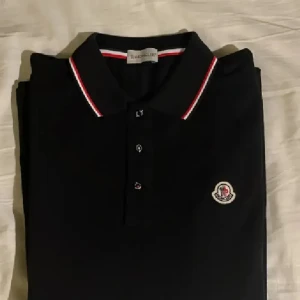 Svart pikétröja från Moncler - Genom swish PostNord!!! Säljer en snygg svart pikétröja från Moncler med klassisk krage och knappar. Den har röda och vita detaljer på kragen och ärmsluten, samt Monclers logga på bröstet. Perfekt för en stilren look! ÄKTA!!